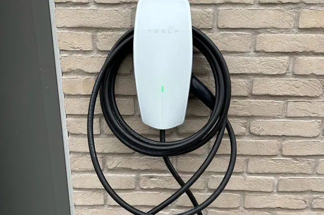 Witte Tesla Wall Connector met opgerolde laadkabel aan een bakstenen muur, geplaatst door Laadnatie.